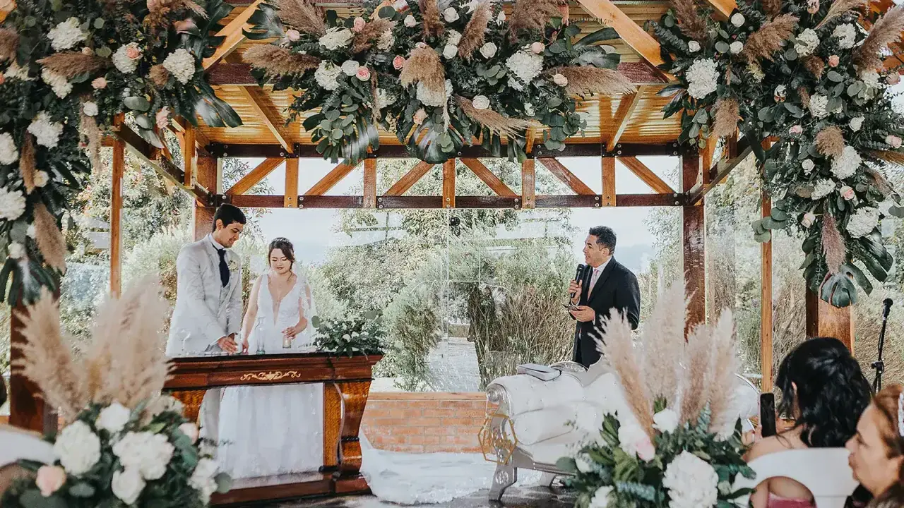 Novios celebran aniversario en altar decorado con flores y pampas. Un oficiante habla mientras la pareja sonríe.