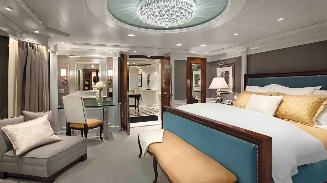 Lussuosa suite crociera con letto king-size, divanetto, lampadario di cristallo e vista su un corridoio elegante.