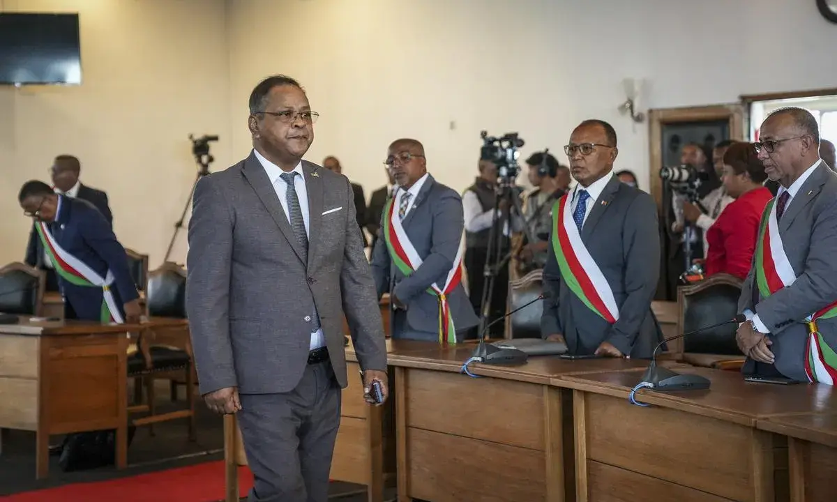Des hommes en costume, certains portant des écharpes aux couleurs nationales, se tiennent dans une salle. L'un d'eux semble être une persona non grata, marchant à l'écart.