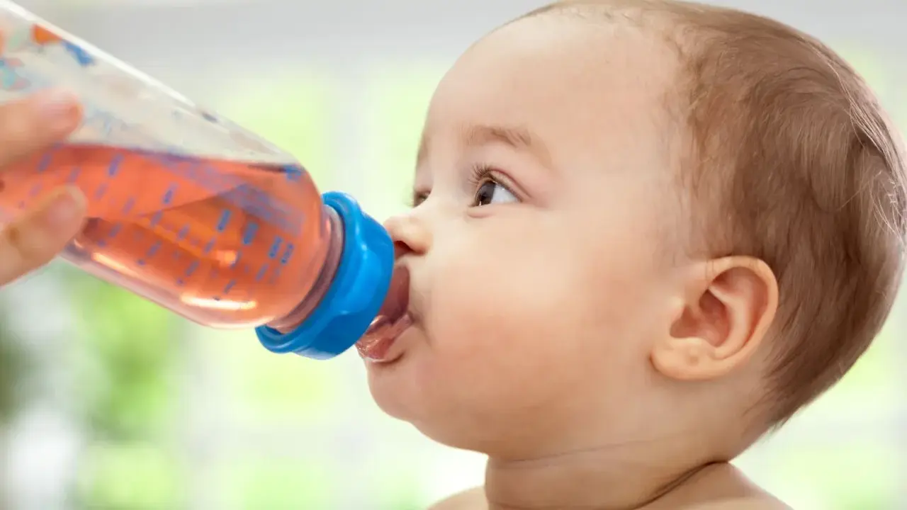 Kleines Baby trinkt aus einer Flasche mit rosafarbenem Tee. Ab wann Tee Baby? Diese Frage stellen sich viele Eltern.