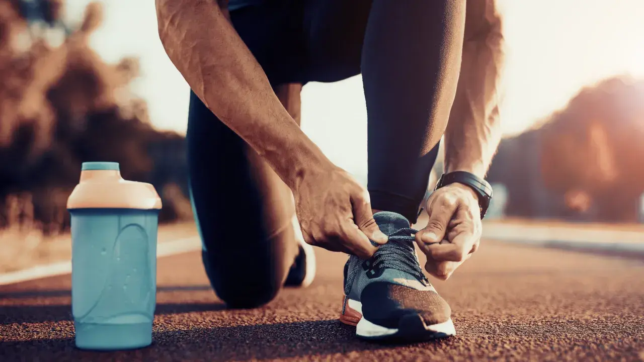 Uomo si allaccia le scarpe da corsa, pronto per correre a stomaco vuoto. Accanto, una borraccia blu.