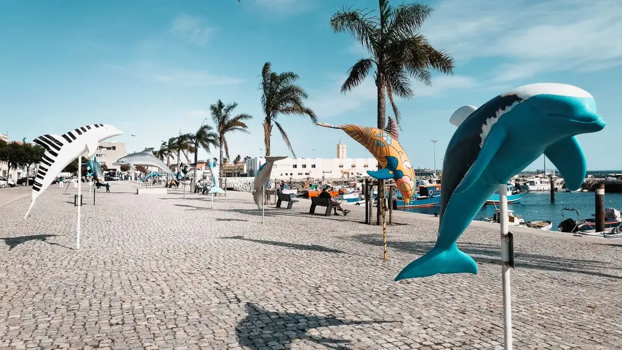 Bunte Delfin-Skulpturen säumen die Promenade in Setúbal, Portugal, unter blauem Himmel und Palmen.
