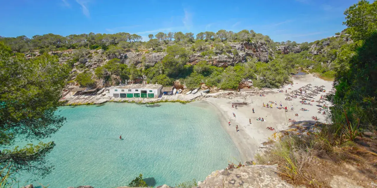 Türkisfarbenes Wasser und ein sonniger Sandstrand in Cala Pi, umgeben von grünen Hügeln. Menschen entspannen sich unter Sonnenschirmen.