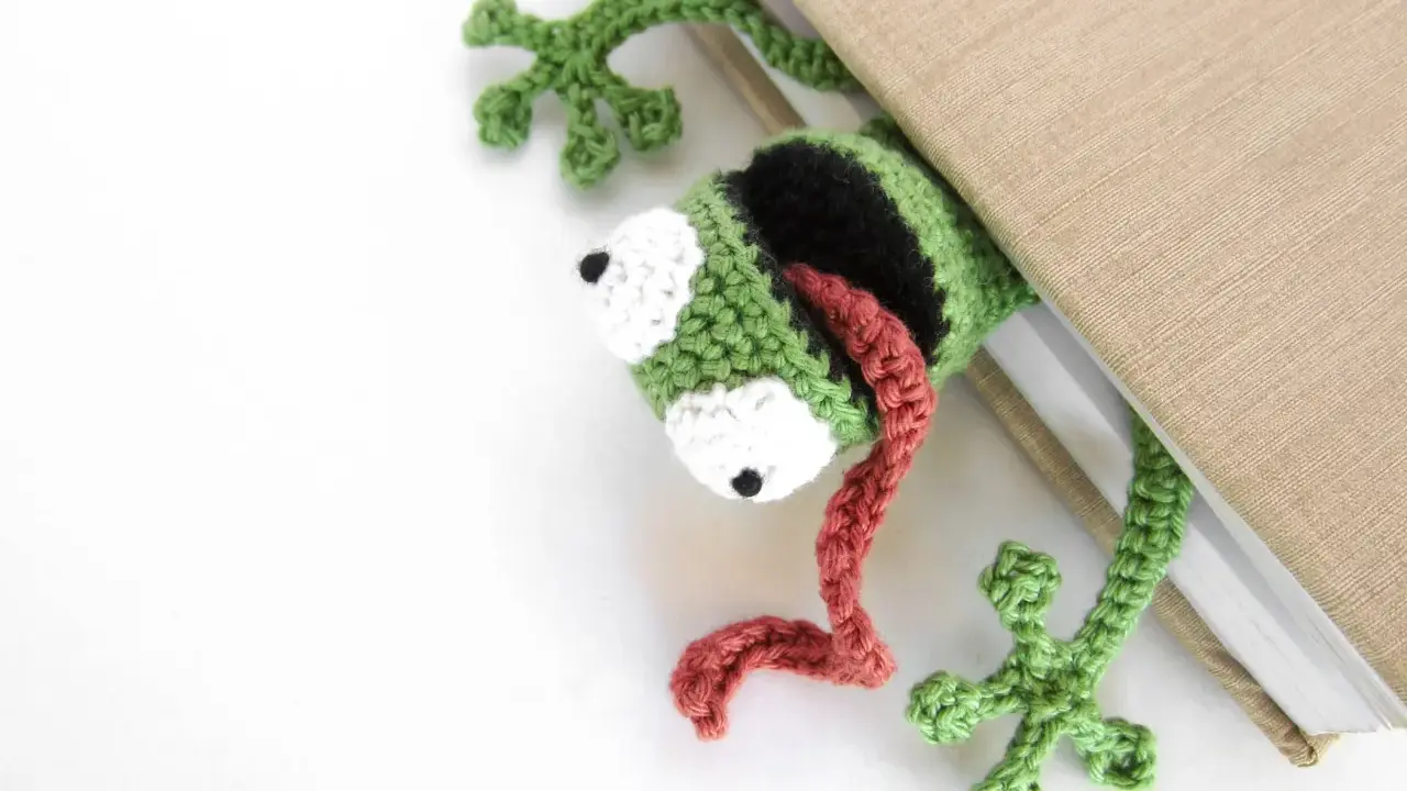 Marcador de libro con ranas amigurumi, verde y negro, con lengua roja, asomando de un libro beige.