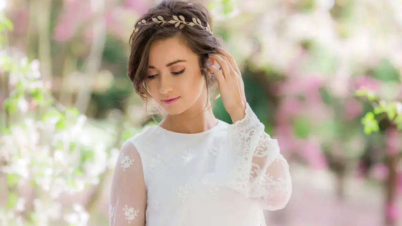 Delikatna tiara na głowę zdobi brunetkę w białej sukni z koronkowymi rękawami, otoczoną kwitnącymi drzewami.