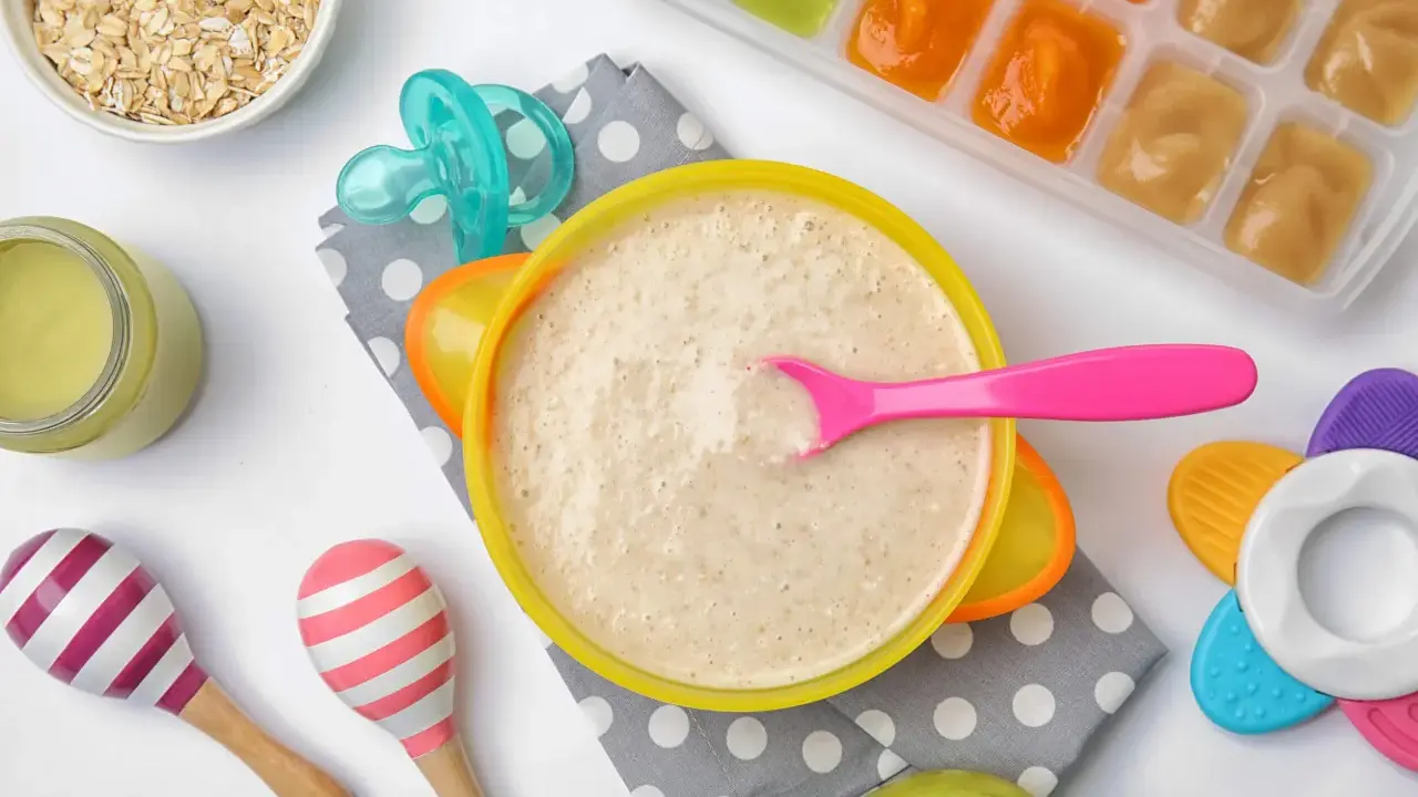Un tazón de papilla de cereales con una cuchara rosa, junto a avena, purés de frutas y juguetes para bebés.