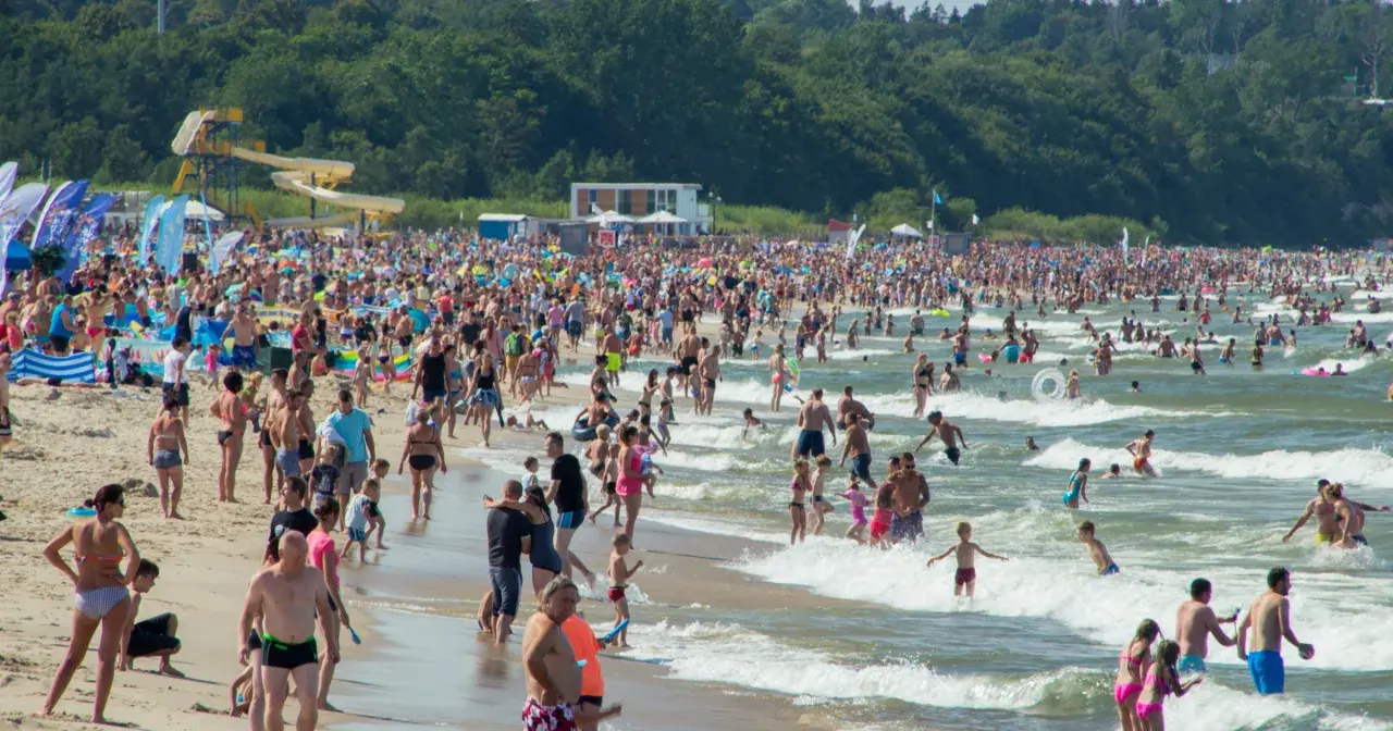 Tłumy ludzi na plaży i w morzu, szukają idealnego miejsca gdzie na wakacje w Polsce. W tle zjeżdżalnie wodne i las.
