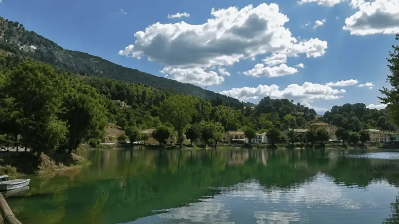 Il lago Sirino, con le sue acque verdi e calme, riflette le case e le colline boscose sotto un cielo azzurro con nuvole bianche.