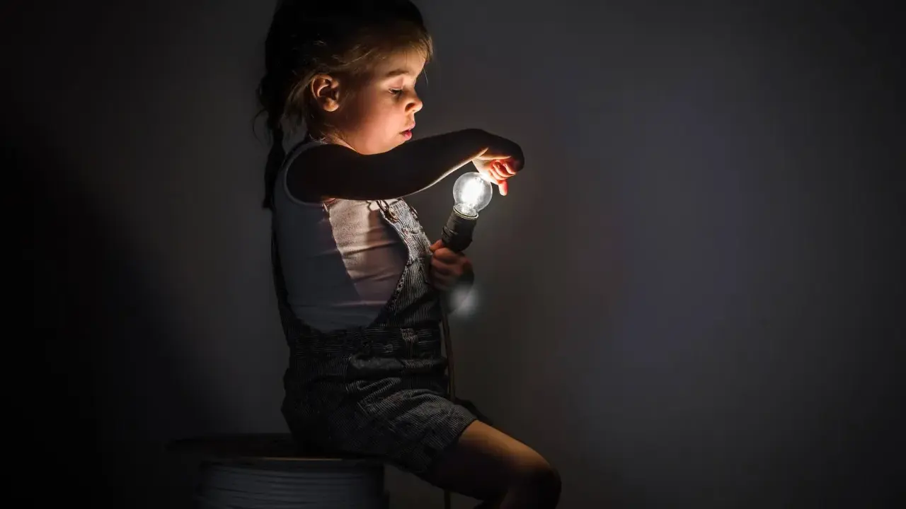 Niña con miedo a la oscuridad, sentada en un rollo de cable, sosteniendo una bombilla encendida.