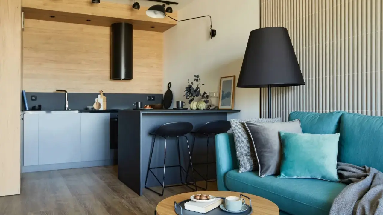 Nowoczesny salon z aneksem kuchennym 22m2. Turkusowa kanapa, czarne hokery przy wyspie, drewniane akcenty i stylowe oświetlenie tworzą przytulną przestrzeń.