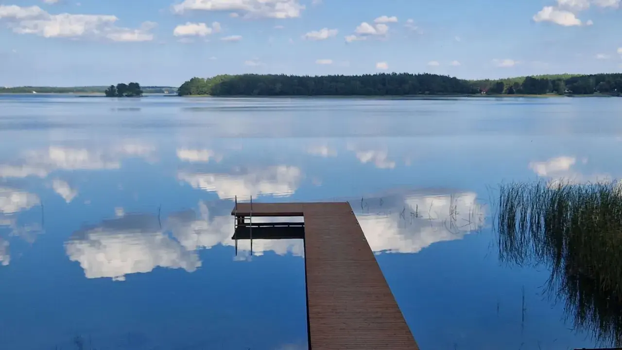Drewniany pomost na spokojnym jeziorze Urszulewskim, odbijającym błękitne niebo z białymi chmurami.