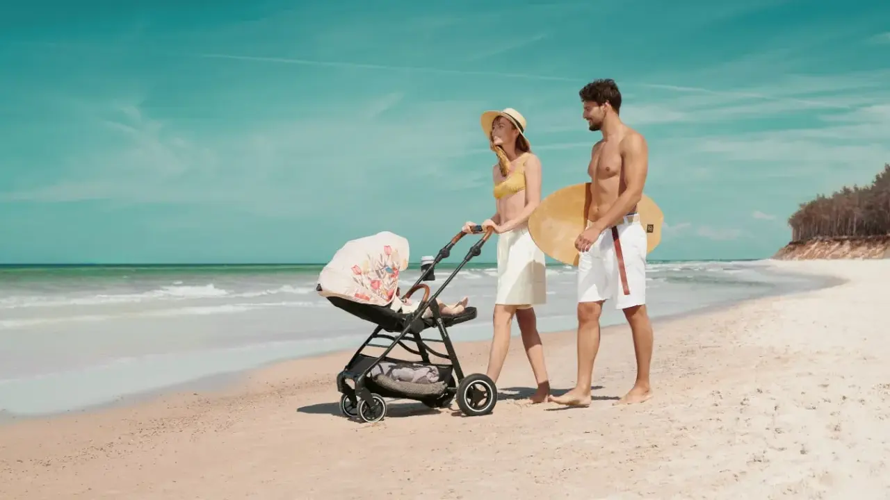 Para spaceruje po plaży z wózkiem Kinderkraft Askoy. Mężczyzna niesie deskę surfingową.