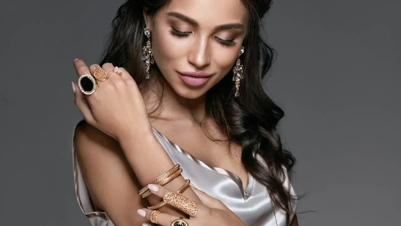 Kobieta w eleganckiej, satynowej sukience prezentuje bogatą biżuterię – złote bransolety, pierścionki z czarnymi kamieniami i kryształowe kolczyki. To kwintesencja glamour stylu ubioru.