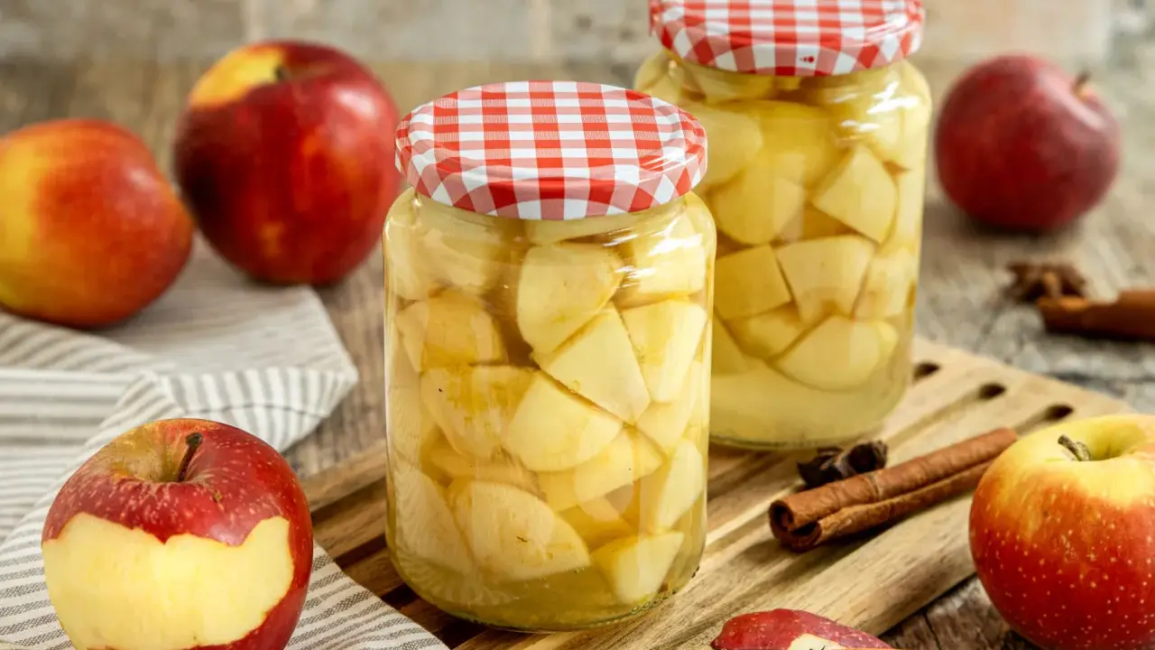 Zwei Gläser mit Apfelstücken, bereit zum Apfel einkochen. Frische Äpfel, Zimtstangen und Sternanis liegen daneben.