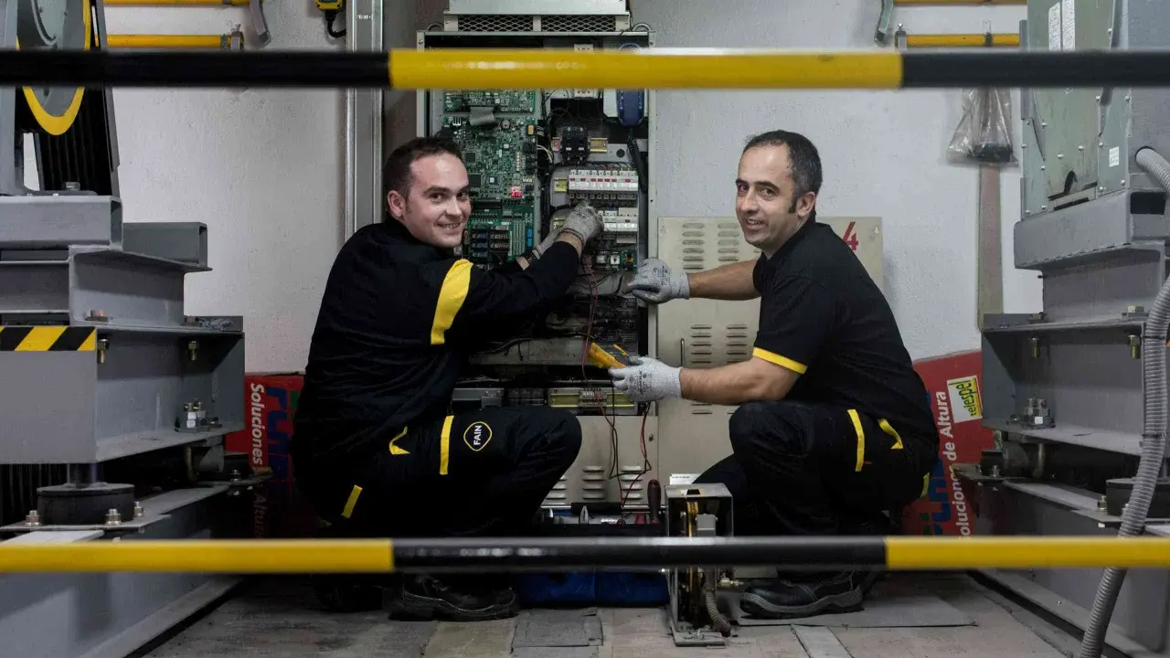 Técnicos realizan mantenimiento de elevadores en un taller, revisando la electrónica y cableado.
