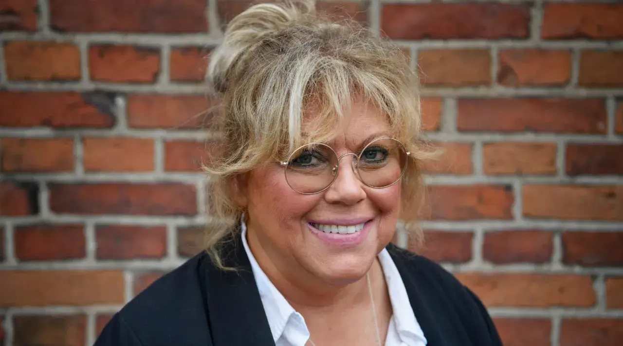 Marie Nilsson Lind ler mot kameran, med sitt blonda hår uppsatt och runda glasögon.
