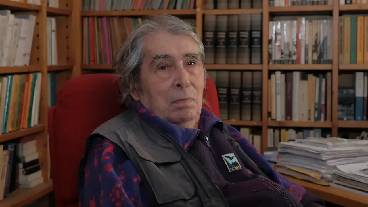 Ferdinando Camon, con i suoi capelli grigi e lo sguardo pensieroso, siede tra scaffali pieni di libri, un ritratto di un intellettuale immerso nel suo mondo.