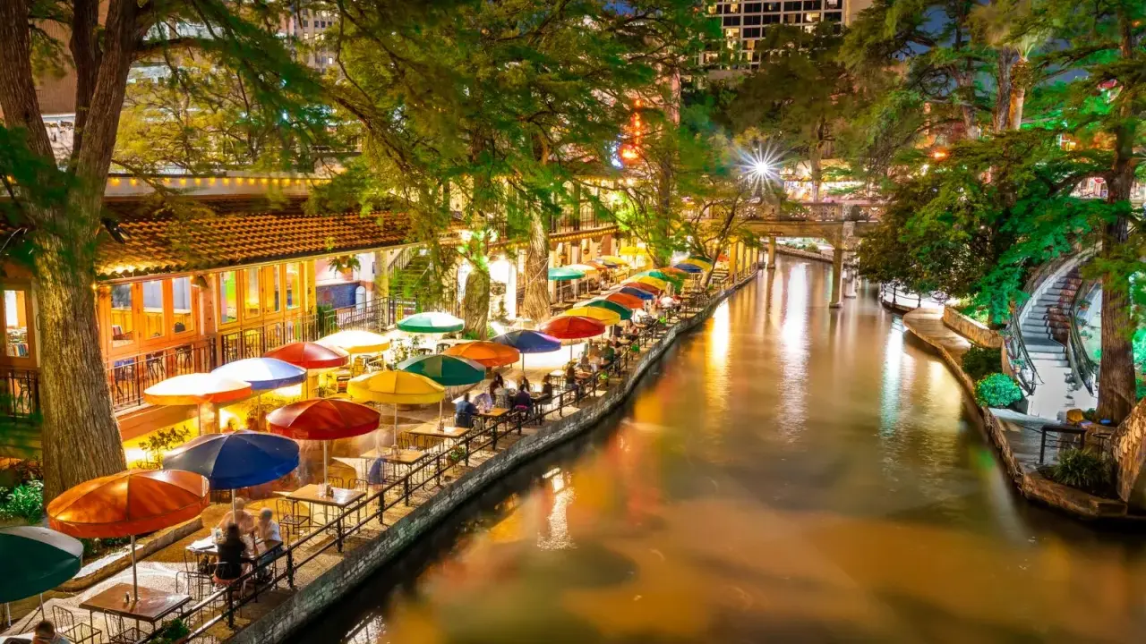 Bunte Sonnenschirme säumen den River Walk in San Antonio, wo Gäste abends in Restaurants am Wasser speisen.