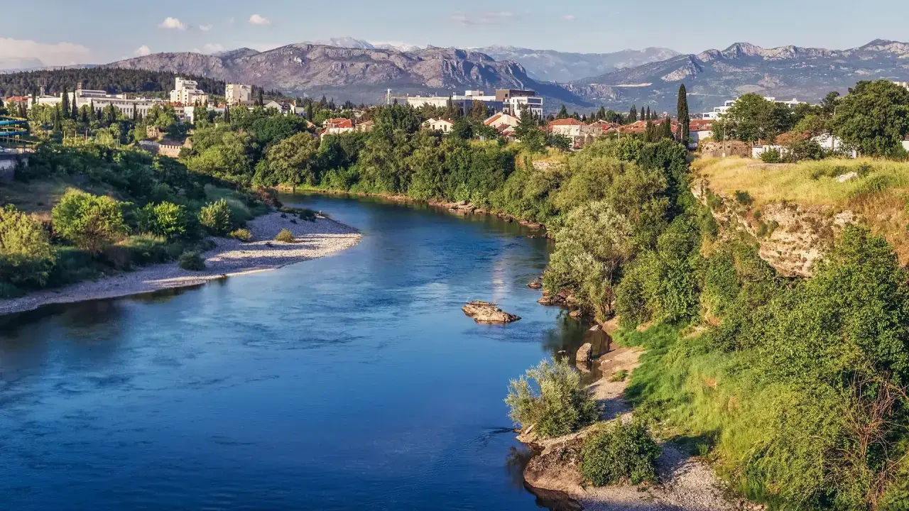 Malowniczy widok na rzekę i miasto Podgorica. W oddali góry, a na pierwszym planie bujna zieleń i kamieniste brzegi. Atrakcje Podgoricy w pełnej krasie.