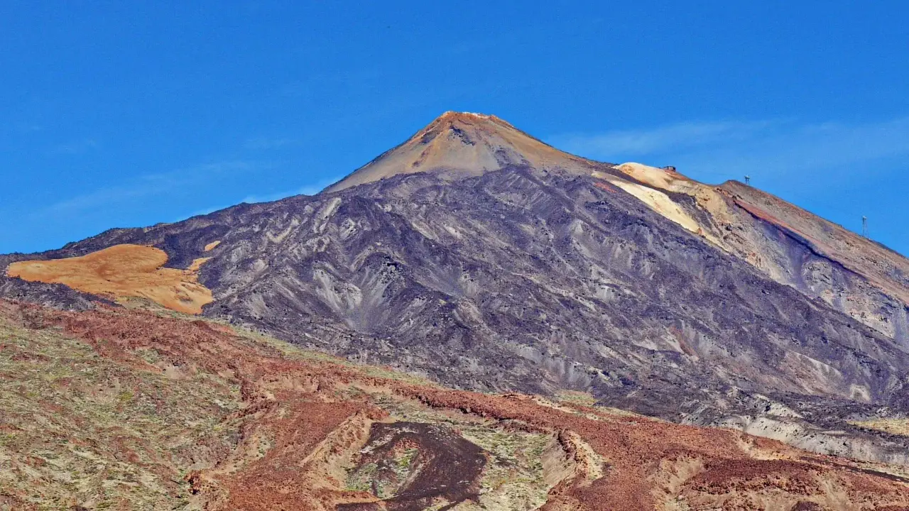Majestätischer Vulkan im Teide Nationalpark unter blauem Himmel. Die Hänge zeigen verschiedene Gesteinsschichten und eine sandfarbene Fläche.