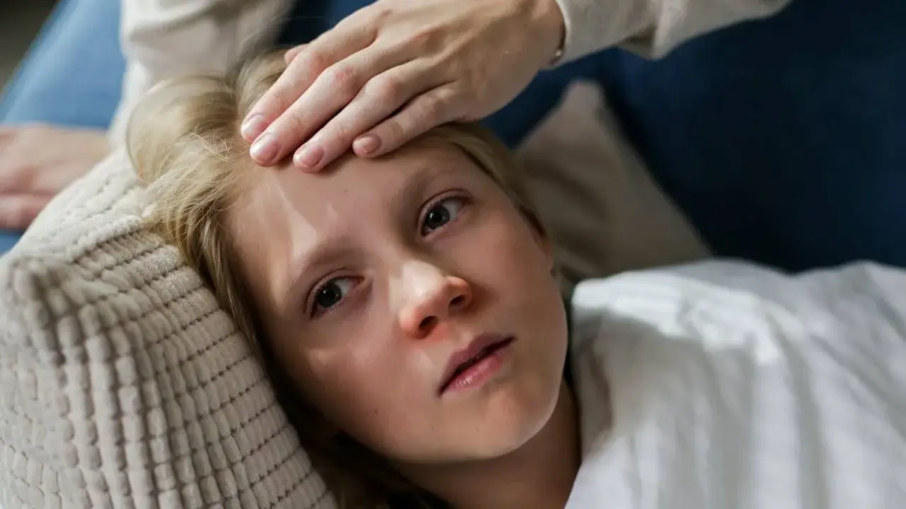 Niña con fiebre, su frente tocada por una mano. Posiblemente un caso de mononucleosis en niños.