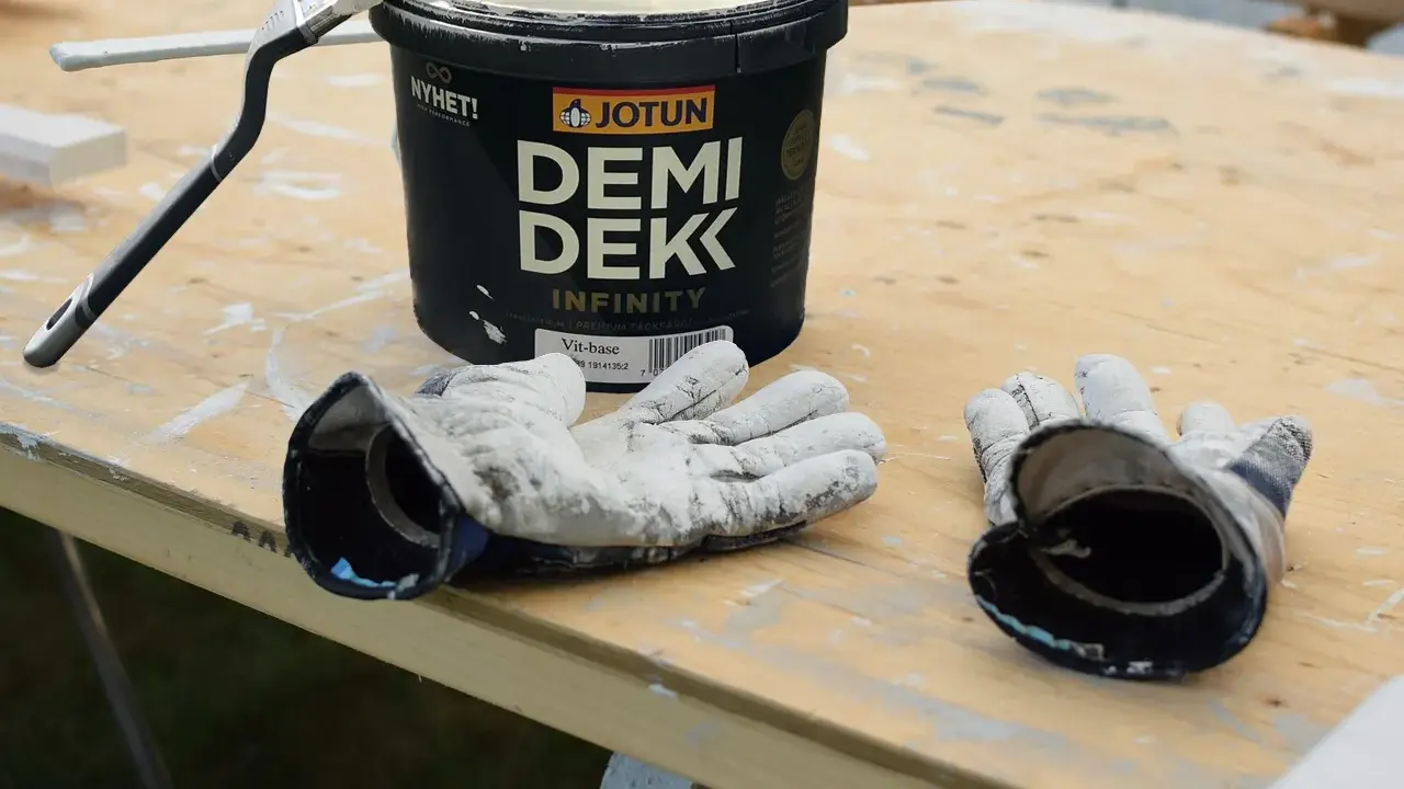 Förberedelser för målning utomhus: en burk Jotun Demi Dekk, pensel och slitna handskar ligger på ett träbord.