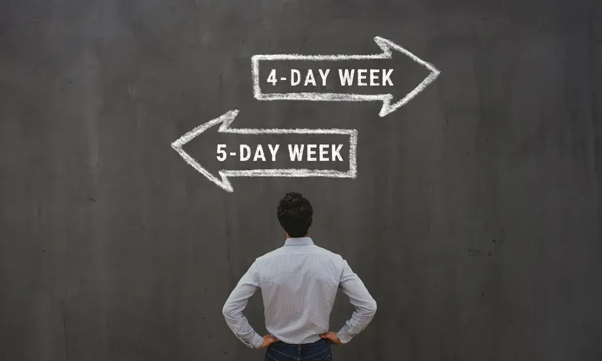 Mężczyzna stoi przed tablicą, na której kredą narysowano dwie strzałki: w prawo "4-DAY WEEK" i w lewo "5-DAY WEEK".