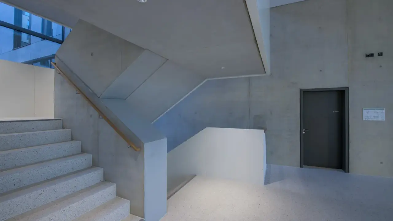 Moderne Treppe mit Sichtbeton-Wänden, die durch die Schalung eine interessante Textur erhalten. Ein dunkler Türrahmen und ein Holzhandschuh ergänzen das Bild.