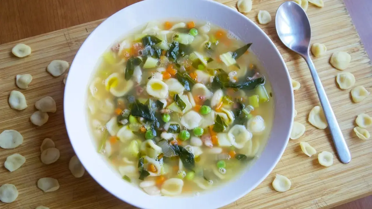 Un reconfortante plato de sopa minestrone, lleno de pasta orecchiette, verduras y frijoles, servido en un tazón blanco sobre una tabla de madera.