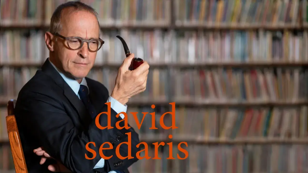 David Sedaris, con pipa in mano, seduto tra scaffali pieni di libri, riflette pensieroso.