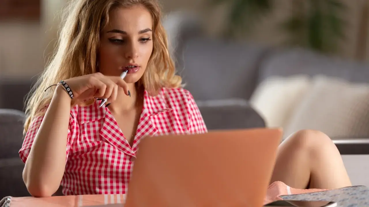 Młoda kobieta w kratkę zastanawia się, jak zacząć rozmowę przez internet, trzymając długopis przy ustach.