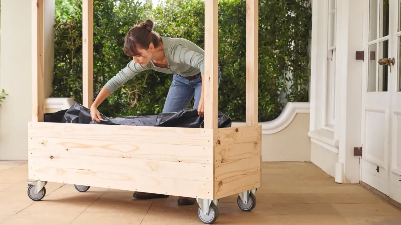 Une femme installe une bâche noire dans une jardinière en bois à roulettes, étape clé pour comment fabriquer toit pour tomates.