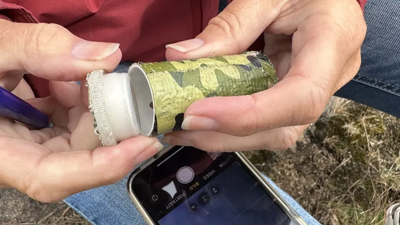 Händer öppnar en kamouflerad behållare, redo för geocaching i Sverige. En mobiltelefon ligger redo att dokumentera fyndet.