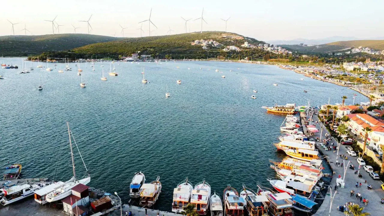 Port w Alacati, Turcja, pełen łodzi i żaglówek, z wiatrakami na wzgórzach w tle.