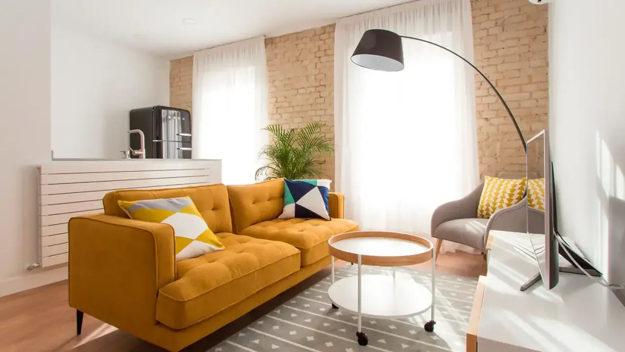 Sofá mostaza con cojines geométricos, mesa auxiliar redonda y alfombra gris. Los colores que combinan con el mostaza resaltan en esta sala.