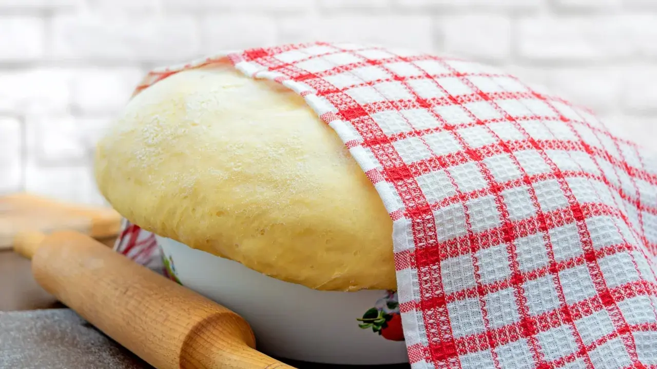 Wyrośnięte ciasto drożdżowe, przykryte ręcznikiem w kratkę, czeka na swój przepis babci. Obok wałek i mąka.
