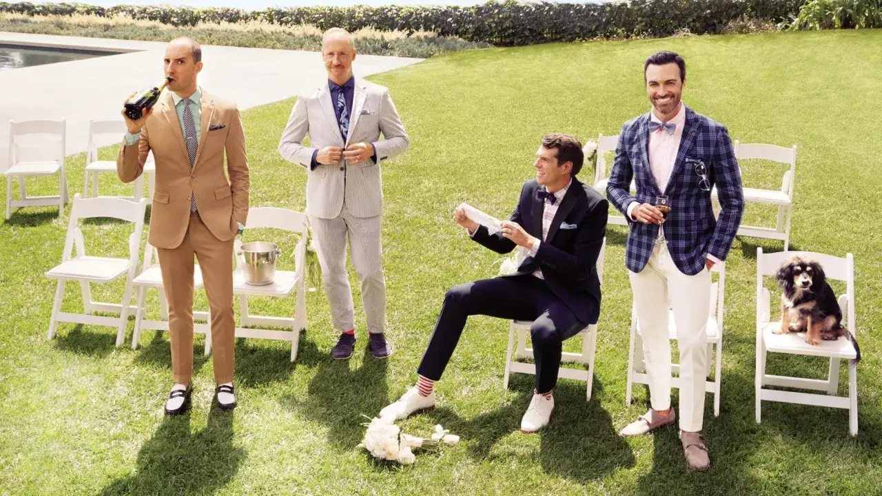 Four men in stylish garden formal wedding attire for male guests pose on a lawn. One drinks champagne, another adjusts his suit, one sits with a bouquet, and the last holds a drink with a dog nearby.