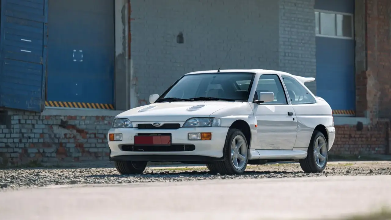 Biały Ford Escort Cosworth z charakterystycznym spojlerem, zaparkowany przed industrialnym budynkiem z niebieskimi drzwiami.