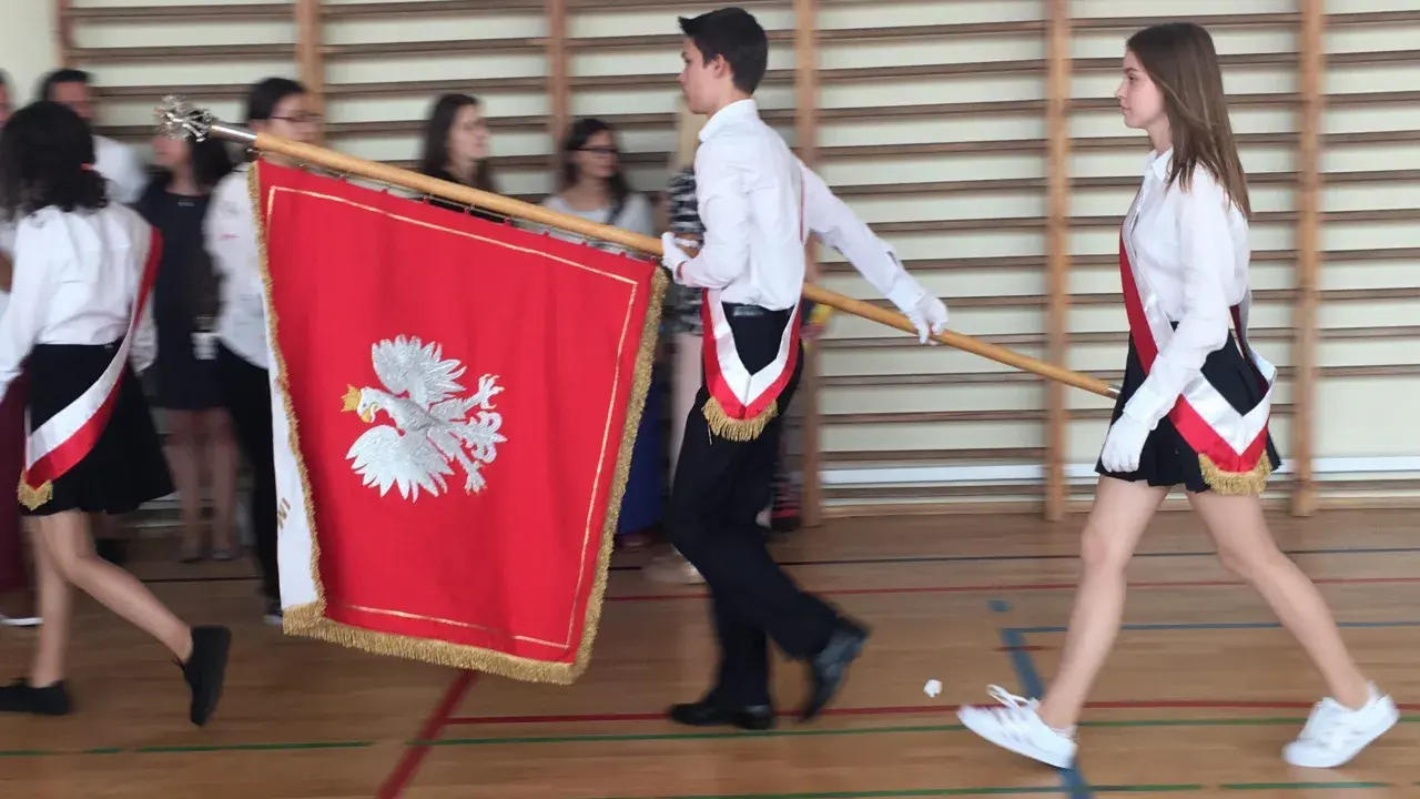 Uczniowie w galowych strojach tworzą poczet sztandarowy, niosąc biało-czerwony sztandar z godłem Polski.