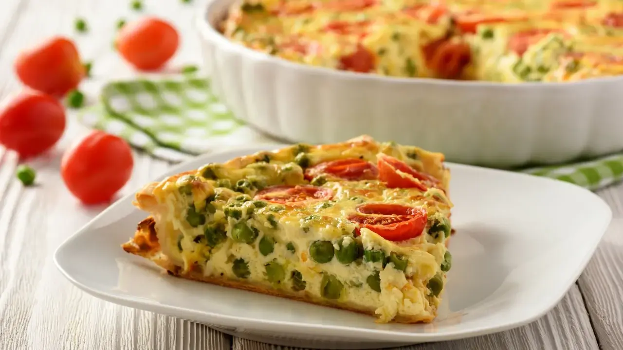 Ein Stück vegetarische Quiche mit Erbsen und Tomaten auf einem Teller. Das ganze Gericht ist im Hintergrund zu sehen.