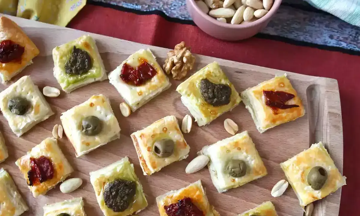 Rustici con pasta sfoglia, piccoli quadrati dorati con olive, pomodori secchi e pesto, disposti su un tagliere di legno.