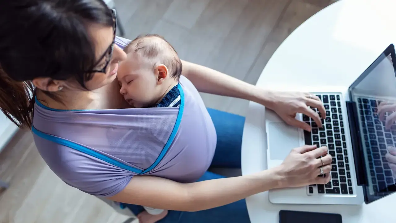 Kobieta w chuście nosi śpiące dziecko i pracuje na laptopie. Czy na macierzyńskim można pracować? Tak, to dowód.
