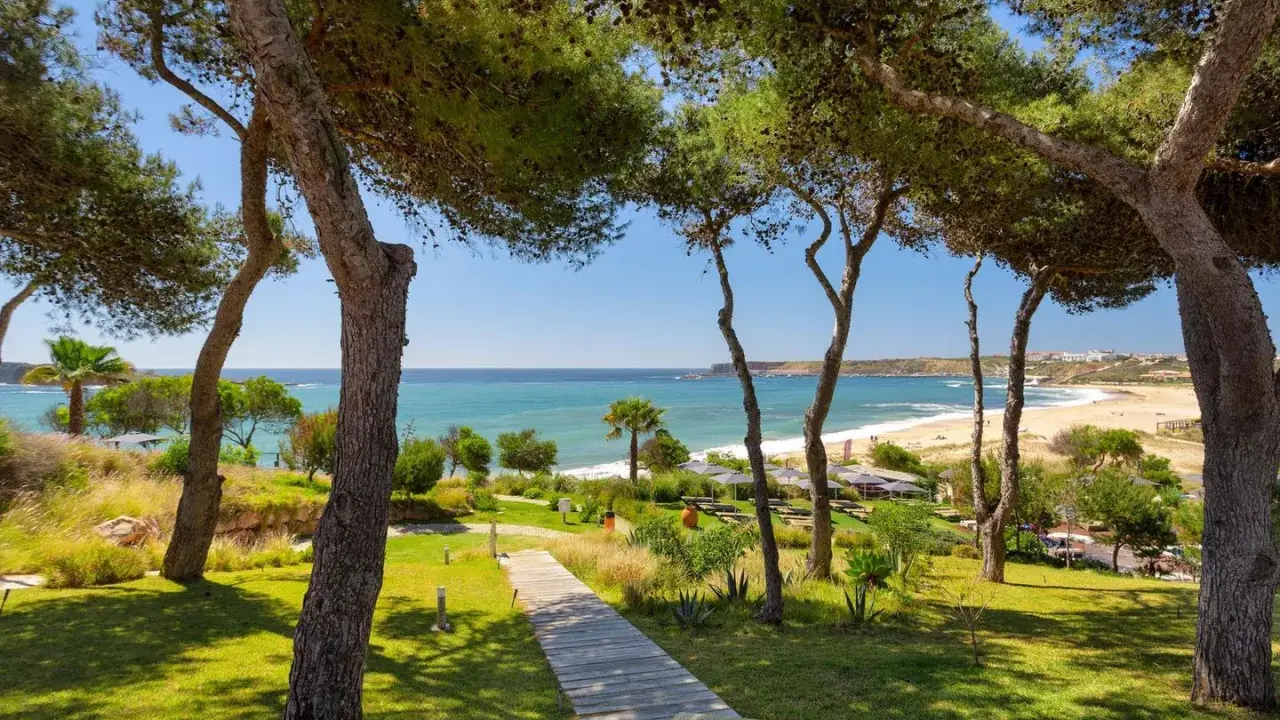 Holzweg führt zu einem sonnigen Strand in Pera, Portugal. Pinienbäume säumen den Weg zum türkisfarbenen Meer.
