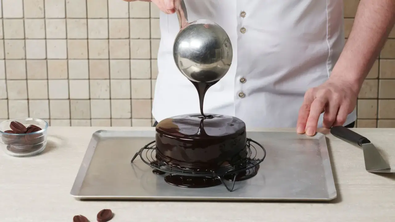 Un chef verse un nappage chocolat brillant sur un gâteau. Des pépites de chocolat sont à côté.