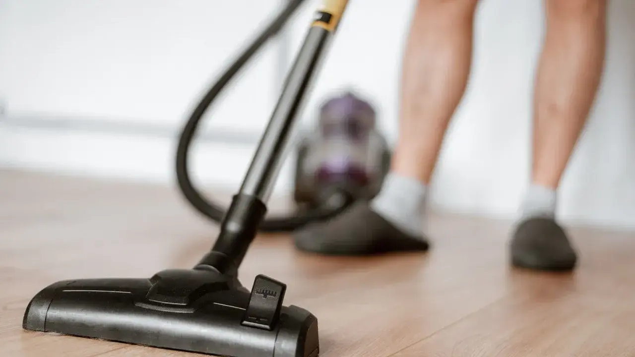 Odkurzacz na drewnianej podłodze, część nóg w kapciach i skarpetkach. To jeden z obowiązków domowych po angielsku – "vacuuming".