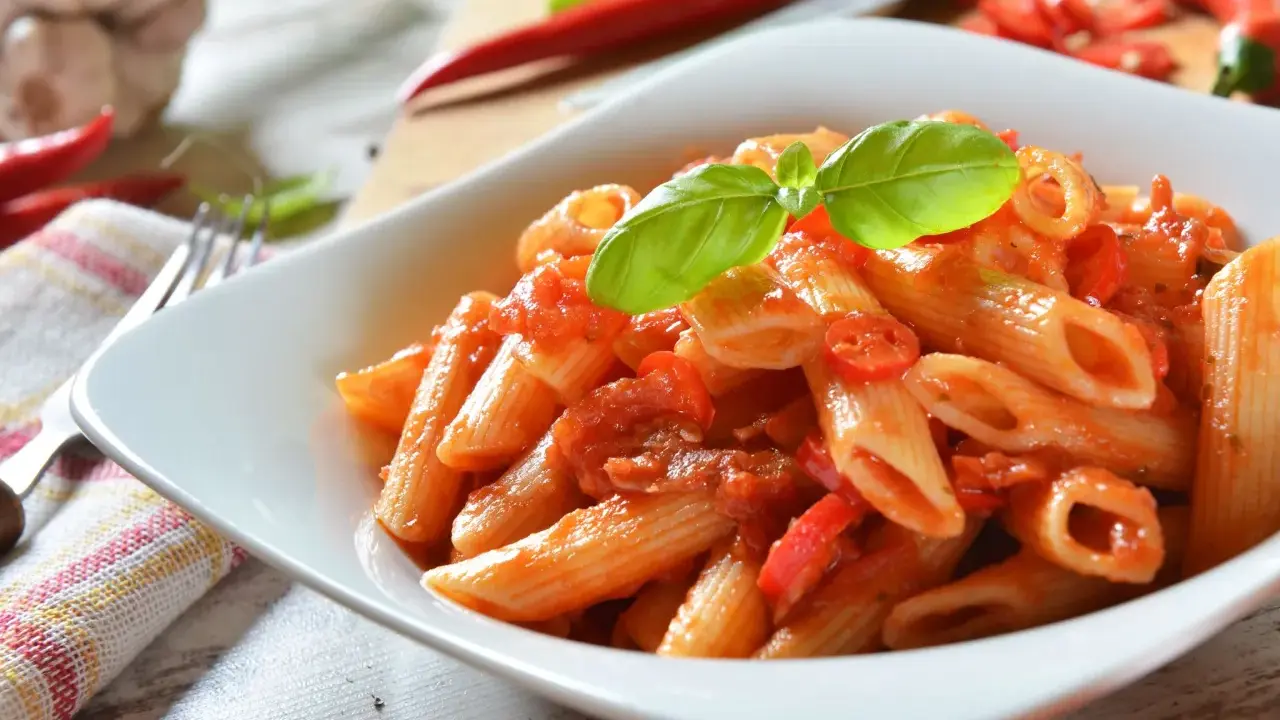 Eine Schüssel Penne Arrabiata, garniert mit Basilikum, mit scharfen Chilis und Knoblauch im Hintergrund.