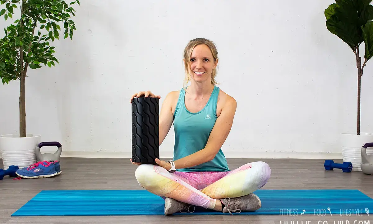 Frau zeigt, wie benutzt man eine Faszienrolle für das Training. Sie sitzt auf einer blauen Matte mit Gewichten und Pflanzen im Hintergrund.