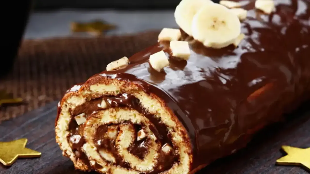 Bûche roulée au chocolat, garnie de crème et recouverte d'un glaçage chocolat beurre brillant, décorée de rondelles de banane et de noisettes concassées.