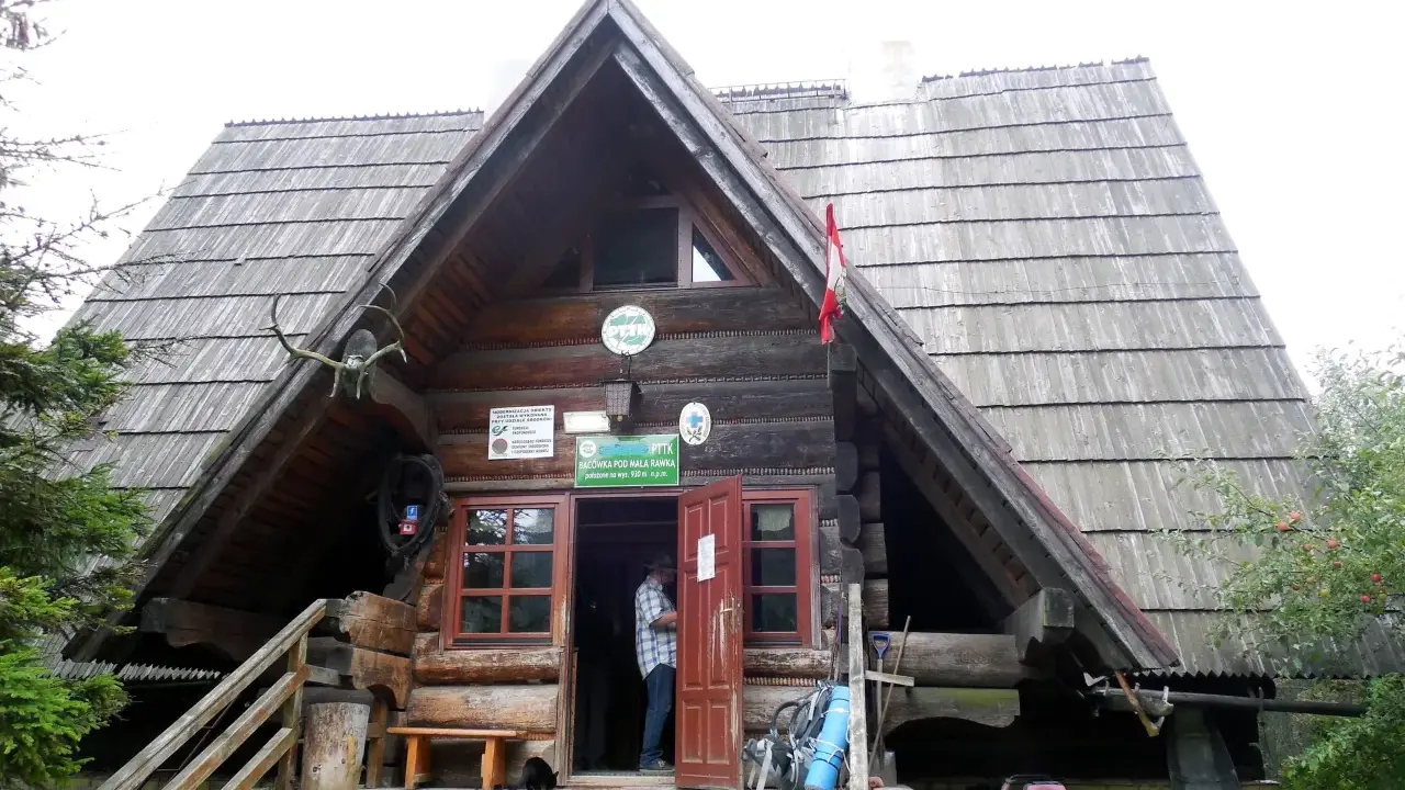 Drewniana bacówka PTTK pod Małą Rawką z otwartymi drzwiami, gdzie stoi mężczyzna. Obok stoją rowery i plecaki.