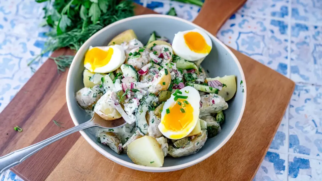 Schüssel mit cremigem Kartoffelsalat mit Gurken, Zwiebeln und gekochten Eiern, garniert mit frischen Kräutern.